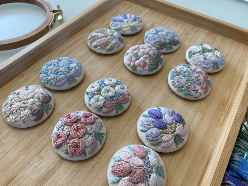 Puede incluir: Diez broches florales bordadas en varios colores y dise&ntilde;os, expuestas sobre una superficie de madera.