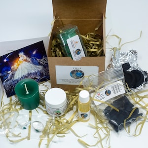 May include: A Yule-themed gift box with a green candle, a jar of pine-scented cream, a bottle of oil, and a small bag of charcoal. The box also contains a small card with an illustration of a woman in a white dress and a starry sky.