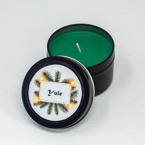 May include: A black tin candle with a green wax top and a white wick. The lid has a festive design with a pine branch, lights, and the word "Yule" on a white tag.
