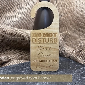 May include: A light-coloured wooden door hanger with a curved top, designed to hang on a bottle. The text "DO NOT DISTURB" is at the top, followed by "Stacey & Gareth ARE MORE THAN Engaged!" in a decorative font. The hanger is made of wood.