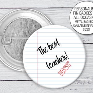 May include: A round pin badge featuring a white background with blue horizontal lines and a vertical pink margin line. The text reads The best teacher! and the word FACT in red outline. A metal pin backing is visible behind the badge.