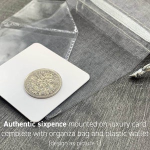 May include: An authentic sixpence coin mounted on a white luxury card, presented with an organza bag and a plastic wallet. The coin is silver-colored and features intricate details. The text "Authentic sixpence mounted on luxury card" is visible.