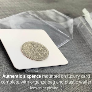 May include: A silver sixpence coin from 1961 is displayed on a white card, accompanied by a clear plastic wallet and a gray organza bag. The text "Authentic sixpence mounted on luxury card complete with organza bag and plastic wallet" is visible.