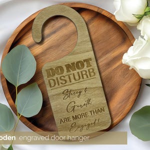 May include: Wooden engraved door hanger with the text "DO NOT DISTURB" and the names "Stacey & Gareth ARE MORE THAN Engaged!". The door hanger is light brown and has a hook at the top. The door hanger is on a wooden tray.