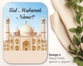 Puede incluir: Una tarjeta azul claro y blanca con una ilustración de una mezquita y el texto "Eid Mubarak Name?" en letra cursiva negra. La tarjeta tiene un borde blanco y un fondo marrón claro. Diseño 2, Acabado brillante, 90 mm x 125 mm.