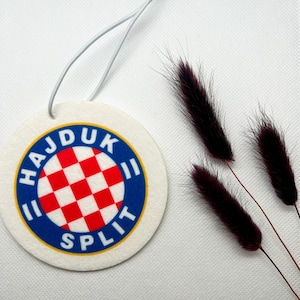 Könnte beinhalten: Ein runder weißer Auto-Lufterfrischer mit einem blauen und gelben Kreis um ein rot-weißes Karomuster. Der Text "HAJDUK SPLIT" ist in einem blauen und gelben Bogen um das Karomuster geschrieben.