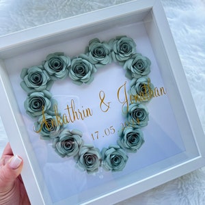 May include: A white frame with a glass front displays a heart shape made of light green paper flowers. The names "Kathrin & Jonathan" and the date "17.05.2021" are written in gold lettering.