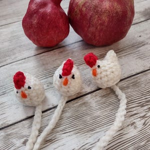 May include: Three white crocheted chickens with red combs and black eyes. The chickens are sitting on a white wooden surface with two red pomegranates in the background.
