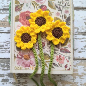 May include: A handmade crochet bookmark featuring three yellow sunflowers with brown centers and green stems. The bookmark is placed on a floral patterned book with a cream-colored cover. The overall aesthetic is rustic and charming.