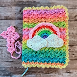 May include: A crocheted rainbow and cloud design book cover with a pink crocheted bookmark. The cover is made with a variety of colors, including pink, orange, yellow, green, blue, and purple.