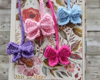 May include: Four crocheted bow bookmarks in different colors: light blue, pink, purple, and hot pink. The bookmarks are attached to a book with floral patterned cover.