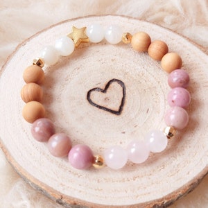 Könnte beinhalten: Ein rosafarbenes, weißes und hölzernes Armband mit einem goldenen Sternanhänger. Das Armband liegt auf einer Holzoberfläche mit einem eingegrabenen Herzen.