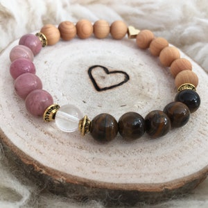 Può includere: Un braccialetto realizzato con perline rosa, marroni e trasparenti. Il braccialetto ha un ciondolo a forma di cuore dorato ed è su una superficie di legno.