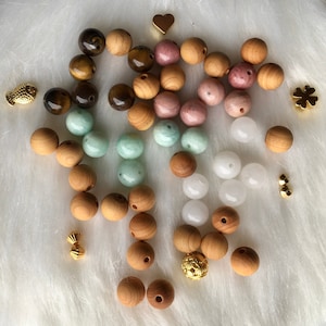 Könnte beinhalten: Eine Auswahl an Perlen in verschiedenen Farben und Materialien, darunter Holz, Jade und rosa Stein. Goldfarbene Charms in Form eines Fisches, Herzens, Kleeblatts und anderer dekorativer Elemente sind zwischen den Perlen verstreut. Die Perlen sind auf einer weißen, flauschigen Oberfläche angeordnet.