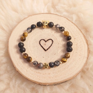 Könnte beinhalten: Ein Armband mit schwarzen Lavasteinen, Tigerauge und grauen Achatperlen. Es enthält auch einen goldenen Geparden-Anhänger und einen goldenen vierblättrigen Kleeblatt-Anhänger.
