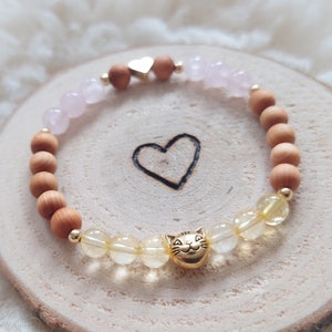 Könnte beinhalten: Ein Armband aus Holzperlen, Rosenquarzperlen und Citrinperlen. Das Armband hat einen goldenen Herzanhänger und einen goldenen Katzenanhänger.
