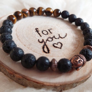 Könnte beinhalten: Ein Armband mit schwarzen Lava-Perlen und Tigerauge-Perlen. Das Armband hat einen Buddha-Anhänger aus Kupfer und die Worte "for you" sind in die Holzoberfläche graviert, auf der es liegt.