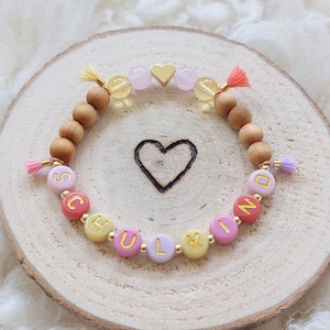 Puede incluir: Un brazalete de cuentas rosa, amarillo y naranja con la palabra "SCHULKIND" escrita en letras doradas. El brazalete tiene un pequeño dije de corazón dorado y cuentas de madera.