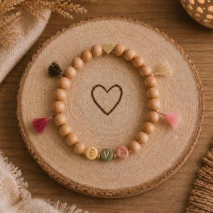 Könnte beinhalten: Ein Perlenarmband mit runden Holzperlen, einem goldfarbenen Herzanhänger oben und drei Buchstabenperlen, die in Gelb, Grün und Rosa EVA buchstabieren. Das Armband ist mit kleinen, farbigen Stoffquasten verziert.