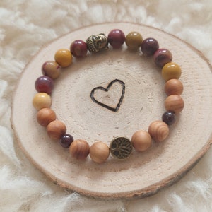 Könnte beinhalten: Ein Holz-Armband mit braunen, gelben und roten Perlen. Das Armband hat einen goldenen Buddha-Anhänger und einen goldenen Baum des Lebens-Anhänger.