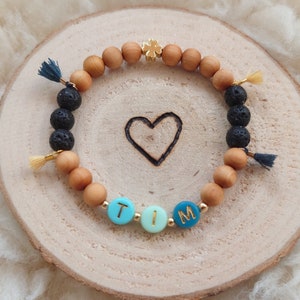 Könnte beinhalten: Ein Holzperlenarmband mit schwarzen Lavasteinen, goldenen Perlen und einem goldenen Glückskleeblatt-Anhänger. Das Armband hat blaue und grüne Perlen, die den Namen "TIM" buchstabieren.