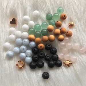 Könnte beinhalten: Sortiment an Perlen in verschiedenen Farben und Materialien, darunter weiß, hellblau, grün, braun, schwarz und rosa. Es gibt auch kleine goldene Charms in Form eines Herzens, eines Sterns, eines Hufeisens und einer Schildkröte.