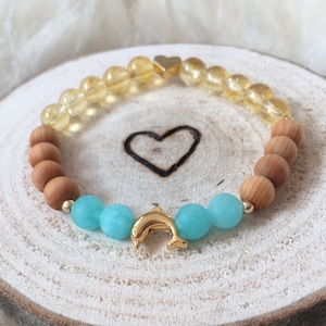 Op de afbeelding: Een kralenarmband met doorschijnende gele kralen, matte bruine houten kralen en helder turquoise kralen. De armband bevat een goudkleurige metalen dolfijnbedel in het midden en een kleine goudkleurige hartvormige kraal aan de bovenkant.