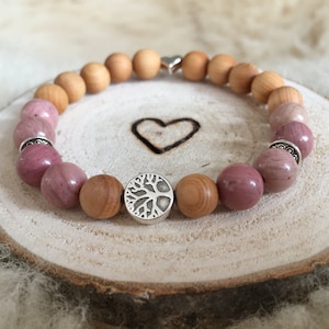 Könnte beinhalten: Ein Armband aus Holzperlen und rosafarbenen Edelsteinperlen. Das Armband hat einen silbernen Anhänger in Form eines Lebensbaums.