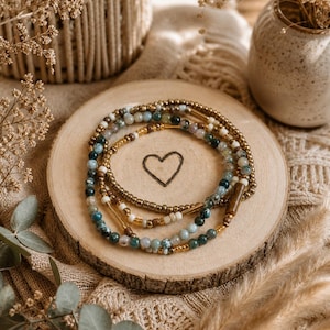 Può includere: Una collezione di braccialetti di perline nei toni del verde, blu, oro e crema, disposti su una fetta di legno con un cuore inciso al centro. I braccialetti sono impreziositi da perline e distanziatori color oro. Lo sfondo include un vaso in macramè e un piccolo vaso con fiori secchi.