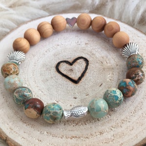 Könnte beinhalten: Ein Armband aus braunen Holzperlen, blauen und braunen Jaspisperlen und silbernen Anhängern in Form eines Herzens, einer Muschel und eines Fisches.