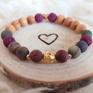 Könnte beinhalten: Ein mehrfarbiges Perlenarmband mit einem goldenen Anhänger. Das Armband besteht aus braunen, violetten und grünen Perlen sowie einem goldenen Herzanhänger.