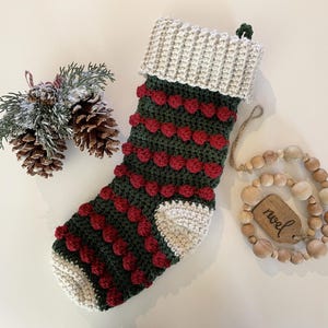 Op de afbeelding: Een groene en rode gehaakte kerstsok met witte pompoentjes en een witte manchet. De sok is versierd met rode en groene strepen. De sok hangt aan een houten kralensnoer met een houten label waarop "kerst" staat.