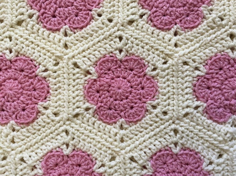 K&ouml;nnte beinhalten: Eine geh&auml;kelte Decke mit wei&szlig;em Hintergrund und rosa Blumenmustern.