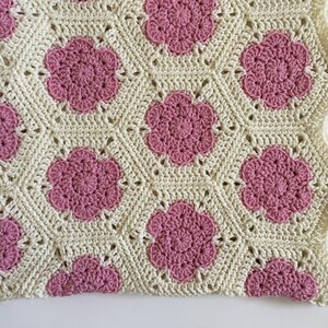 K&ouml;nnte beinhalten: Eine geh&auml;kelte Babydecke mit wei&szlig;em Hintergrund und rosa Blumenmustern.
