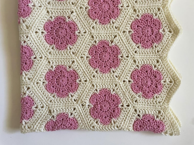 K&ouml;nnte beinhalten: Eine geh&auml;kelte Babydecke mit wei&szlig;em Hintergrund und rosa Blumenmustern.