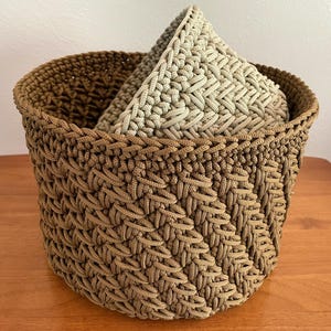 May include: A brown crocheted basket with a beige crocheted pillow inside. The basket is made with a woven pattern and has a round shape.