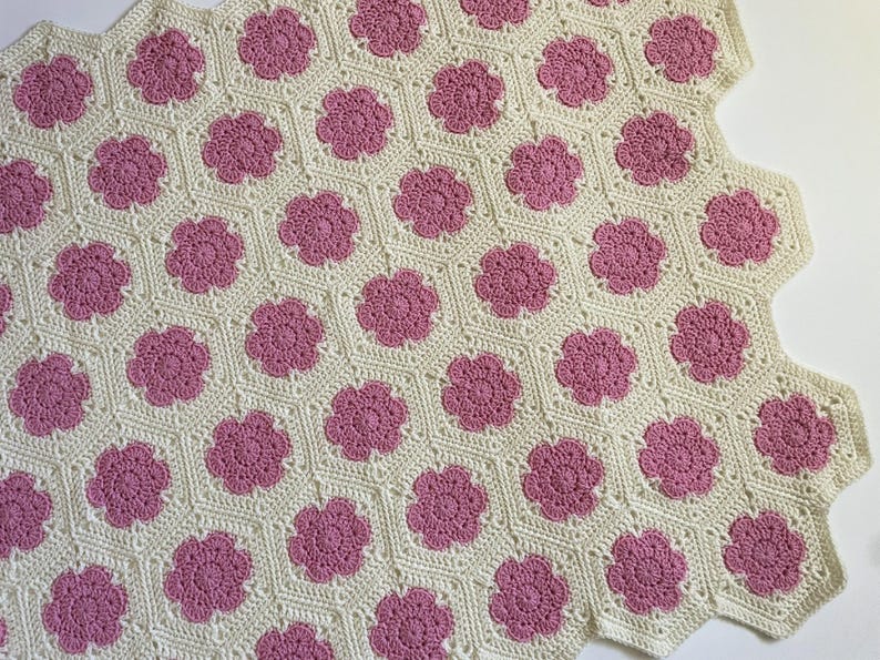 K&ouml;nnte beinhalten: Eine geh&auml;kelte Decke mit wei&szlig;em Hintergrund und rosa Blumenmustern.