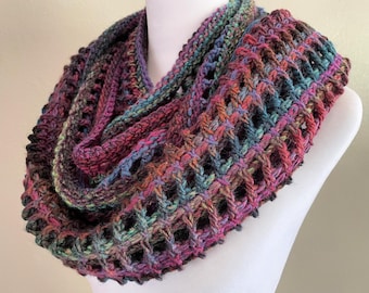 May include: A colourful crocheted infinity scarf with a lacy pattern. The scarf is made with a variety of shades of purple, blue, green, and brown yarn.