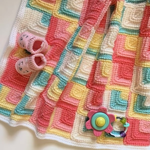 Puede incluir: Una manta de bebé de ganchillo en tonos de rosa, amarillo, blanco y azul. La manta está hecha con un punto texturizado y tiene un borde blanco. Un juguete para bebé está sobre la manta.