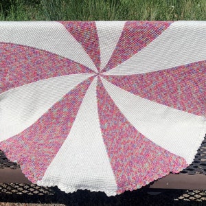 May include: A white and pink crocheted baby blanket with a spiral design. The blanket has a white crocheted border.