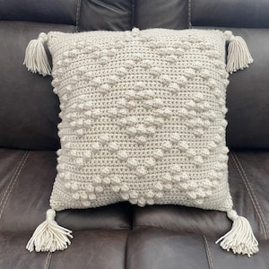 Puede incluir: Un cojín cuadrado de ganchillo gris claro con un patrón de puntos en relieve texturizado. El cojín tiene cuatro borlas, una en cada esquina. El cojín está sobre un sofá de cuero marrón.