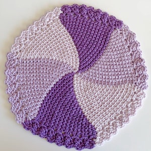 May include: A purple and light purple crochet coaster with a scalloped edge. The coaster is made with a Tunisian crochet stitch.