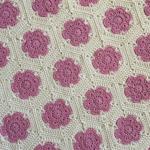 May include: A crocheted blanket with a repeating pattern of pink flowers on a white background.