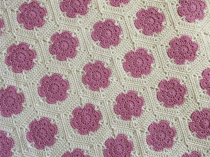 K&ouml;nnte beinhalten: Eine geh&auml;kelte Decke mit einem sich wiederholenden Muster aus rosa Blumen auf wei&szlig;em Hintergrund.