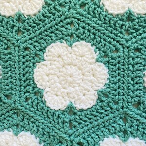 K&ouml;nnte beinhalten: Eine geh&auml;kelte Decke mit einem sich wiederholenden Muster aus wei&szlig;en Blumen auf einem t&uuml;rkisfarbenen Hintergrund.