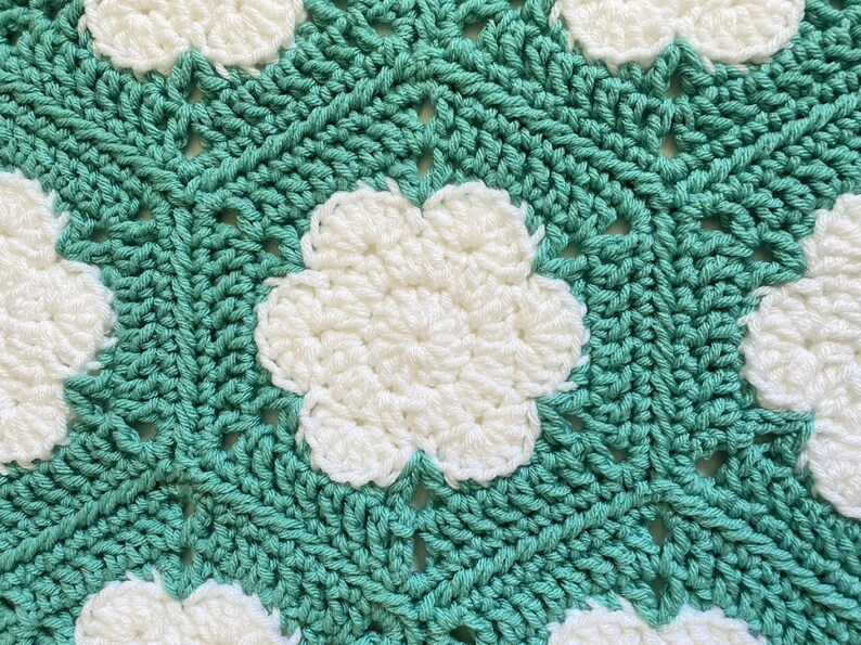K&ouml;nnte beinhalten: Eine geh&auml;kelte Decke mit einem sich wiederholenden Muster aus wei&szlig;en Blumen auf einem t&uuml;rkisfarbenen Hintergrund.