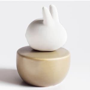 May include: A white ceramic bunny figurine sits on top of a round, light brown ceramic base.