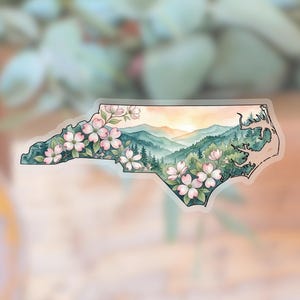 May include: A watercolour sticker in the shape of North Carolina, depicting pink dogwood blossoms, green mountains, and a sunset. The sticker has a transparent border and a soft, blurred background.