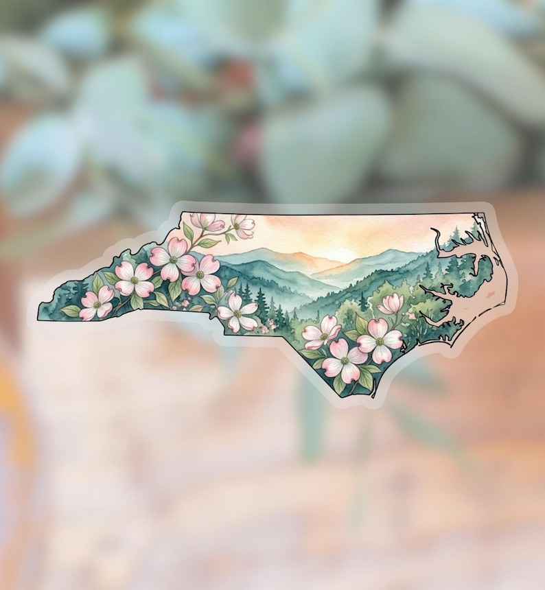 May include: A watercolour sticker in the shape of North Carolina, depicting pink dogwood blossoms, green mountains, and a sunset. The sticker has a transparent border and a soft, blurred background.