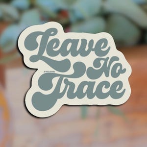 May include: A sticker with the words "Leave No Trace" in a retro font. The text is a muted teal color, outlined in off-white, and set against a blurred background. The sticker also includes the text "BY WILD NATURE".